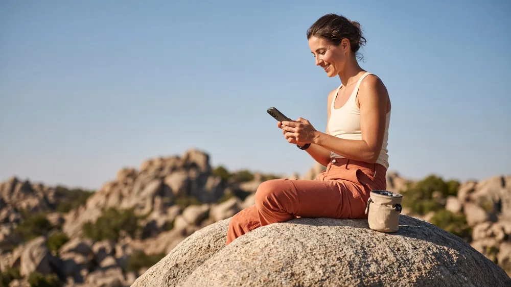 Mujer sentada sobre una gran roca en un paisaje soleado tras escalar sonríe mirando el teléfono
